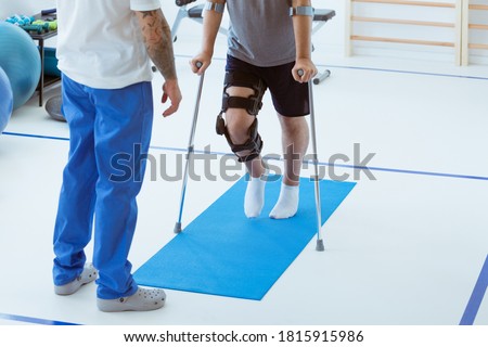 Man after car accident in an orthosis and on crutches learning to walk in the clinic, helpful therapist near him Royalty-Free Stock Photo #1815915986