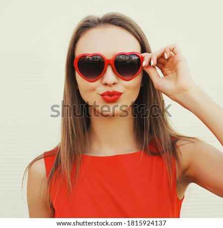 Close up portrait of attractive young woman blowing red lips sending sweet air kiss wearing a heart shaped sunglasses on white background