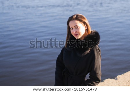 Young, urban woman, brunette in a coat on the shore. In the background is the river. European woman, 27 years old 