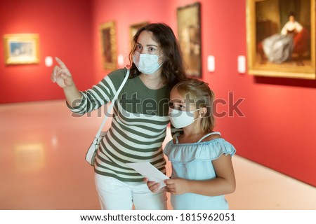 Girl with woman in masks looking with interest at painting in the museum, using guidebook