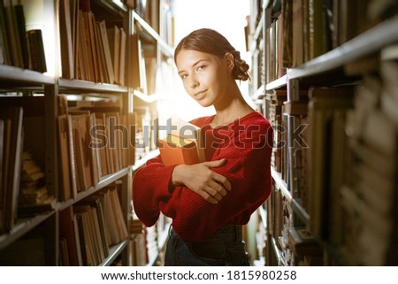 A young girl, dressed in a red sweater, holds a book in her hands against the background of the library. Education concept.