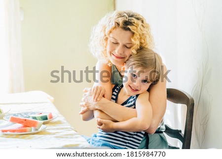 Lovely mother hugging her cute four years old son are smiling while having a breakfast in kitchen. Bright morning in the kitchen. Concept of caressing and love.