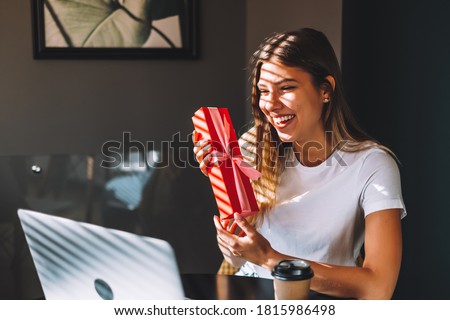 Happy young woman opening gift in front of laptop during video call or chat, celebrating birthday online. Concept of distance relations, celebrations and lifestyle.