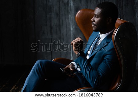 African American businessman in a blue classic suit sits in an aviator loft armchair with a phone in his hands. Royalty-Free Stock Photo #1816043789