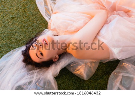 top view of young brunette woman wrapped in polyethylene lying on grass, ecology concept