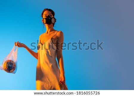 brunette woman in gas mask holding plastic bag with globe on blue, ecology concept