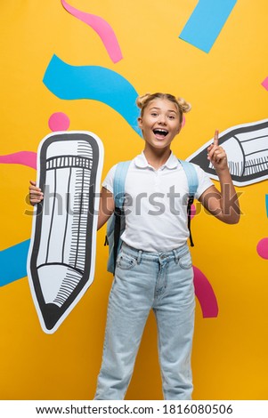 Excited schoolgirl having idea while holding paper art on yellow background