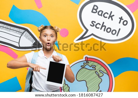 Surprised child pointing with finger at digital tablet near speech bubble and paper art on yellow background