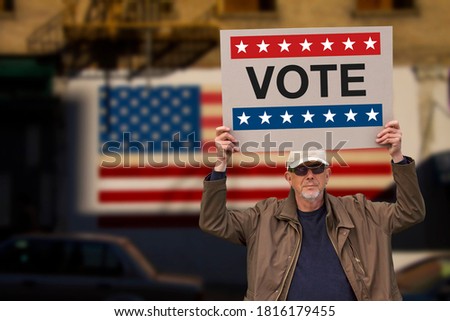 Man with cap blue jeans and  sunglasses holding a cardboard sign with text VOTE above his head with american stars and stripes flag on a wall in the background. American Presidential election image.