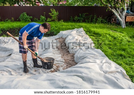 A man pours sand onto a geotextile outside. Garden work, do-it-yourself pool