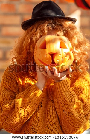 Young woman with red hair wearing hat standing at vacation home outdoors holding jack-o'-lantern on face preparing for halloween posing to camera playful close-up