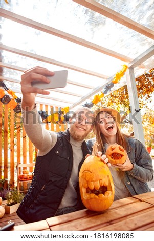 Young couple man and woman sitting at table outdoors at vacation home making jack-o'-lantern preparing for halloween taking selfie photo on smartphone posing with carved pumpkins to camera laughing