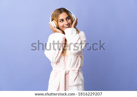 Young blonde Uruguayan woman isolated on blue background in pajamas and holding a pillow and listening music