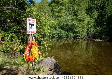 Portage entrance sign for canoeists on a post with an orange lifesaving ring buoy by a brown river in a green forest.