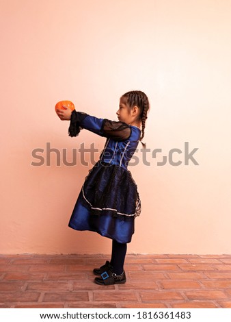little girl disguised in a blue witch costume, in front of a pink background, holding a pumpkin