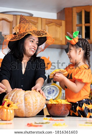 A woman with a kid wearing customes and carving a pumpkin at a decorated kitchen in Halloween