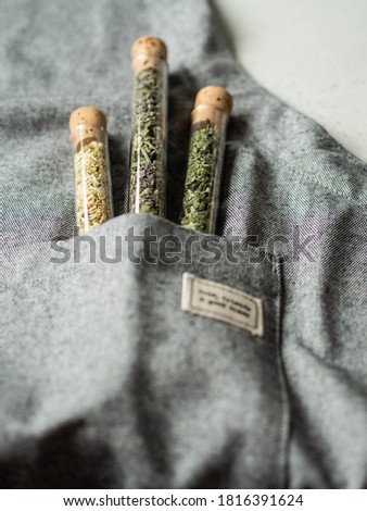glass test tubes with dry green herbal ingredients in pocket of a gray apron. Selective focus.