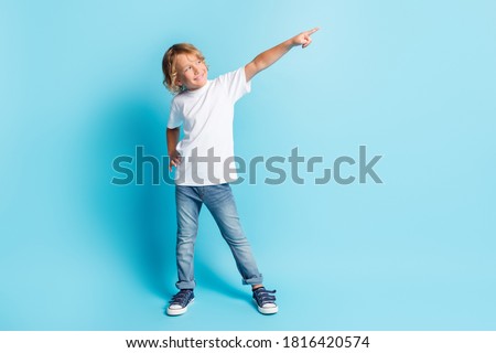 Photo of little kid raise hand indicate finger look empty space wear white shirt jeans sneakers isolated blue color background