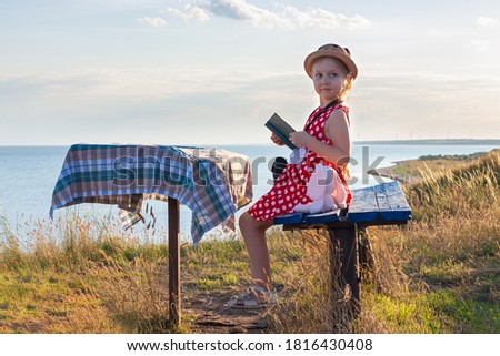 Child girl in hat and polka-dot dress sitting on vintage bench reading a book. Kid looking at Holy Bible in hands and praying on sea lanscape background. Friendship peace religion faith hope concept.