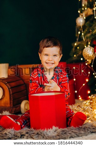 Little boy opening the box present, weared in Christmas pajama sitting in front of New Year pine-tree