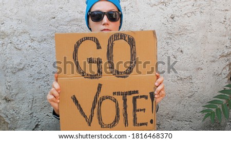 Woman shows cardboard with Go Vote sign on brick wall urban background. Voting concept. Make the political choice, use your voice. Young lady in blue hat invite to go to the presidential elections.
