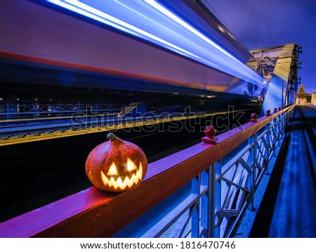 Halloween pumpkin with a glowing grimace at night on the railway bridge. In the background there are rails and blurry streaks of lights from a speeding train. Mystical picture of Halloween in the city