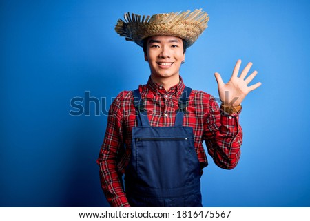 Young handsome chinese farmer man wearing apron and straw hat over blue background showing and pointing up with fingers number five while smiling confident and happy.