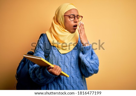 Young African American student woman wearing muslim hijab and backpack holding book bored yawning tired covering mouth with hand. Restless and sleepiness.