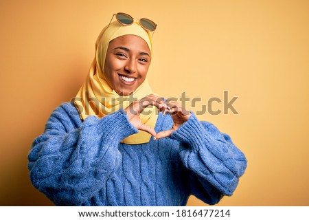 Young African American student woman wearing yellow muslim hijab and sunglasses smiling in love showing heart symbol and shape with hands. Romantic concept.