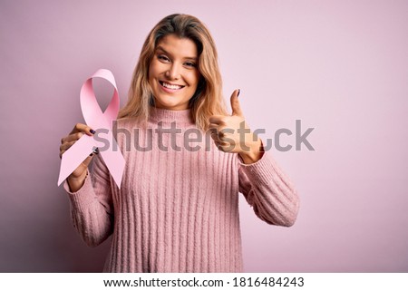 Young beautiful blonde woman asking for support cancer holding pink ribbon symbol happy with big smile doing ok sign, thumb up with fingers, excellent sign