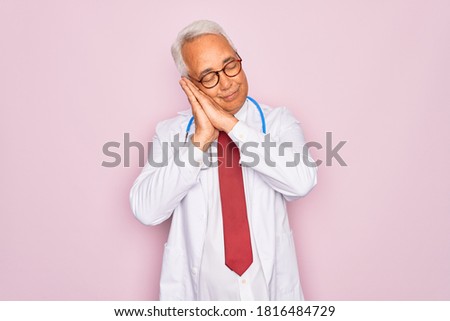 Middle age senior grey-haired doctor man wearing stethoscope and professional medical coat sleeping tired dreaming and posing with hands together while smiling with closed eyes.