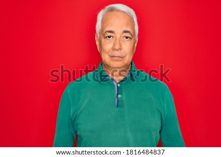 Middle age senior grey-haired man wearing casual sweater over red isoalted background with serious expression on face. Simple and natural looking at the camera.