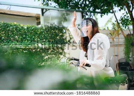 Young asian woman wearing white raincoat and face shield say hi to friend on bicycle.
