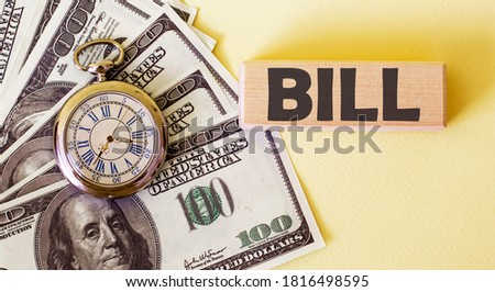 The word Bill is written on a wooden block, a stack of 100 dollars and a clock on a yellow background. American currency. Money and financial concept.