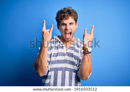 Young blond handsome man with curly hair wearing casual striped sweatshirt shouting with crazy expression doing rock symbol with hands up. Music star. Heavy concept.