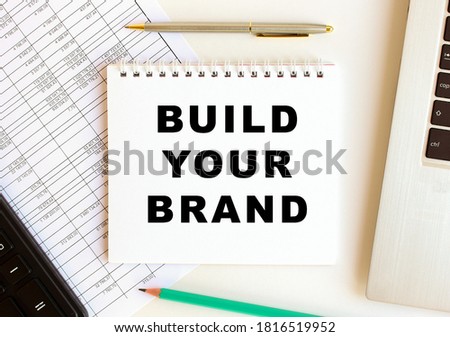Notepad with text BUILD YOUR BRAND on a white background, near laptop, calculator and office supplies. Business concept.