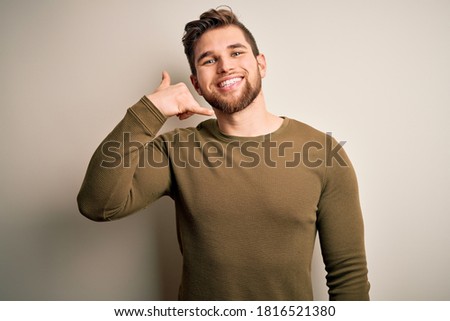 Young blond man with beard and blue eyes wearing green sweater over white background smiling doing phone gesture with hand and fingers like talking on the telephone. Communicating concepts.