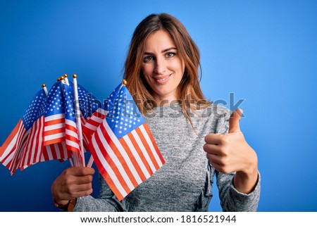 Young beautiful patriotic woman holding united states flags celebrating inependence day happy with big smile doing ok sign, thumb up with fingers, excellent sign