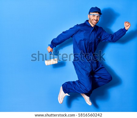 Young handsome hispanic man wearing painter uniform and cap smiling happy. Jumping with smile on face holding roller doing winner sign with fists up over isolated blue background