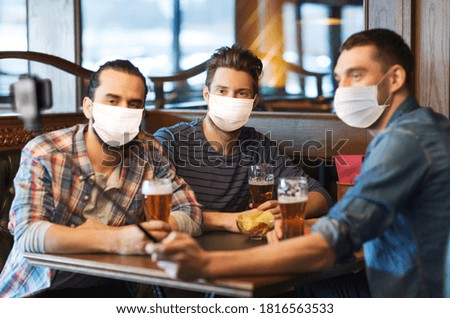 male friendship, leisure and pandemic concept - men or friends in face protective medical masks for protection from virus drinking beer and taking picture with smartphone selfie stick at bar or pub