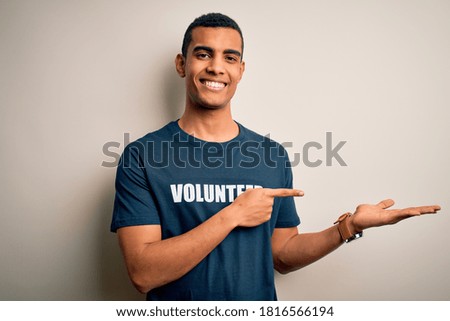 Young handsome african american man volunteering wearing t-shirt with volunteer message amazed and smiling to the camera while presenting with hand and pointing with finger.