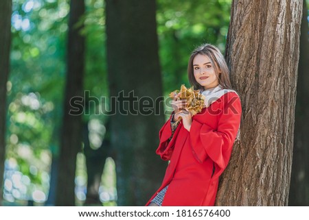 Portrait of young girl in the autumn weather in warm clothes near tree, holding a bundle of leaves.