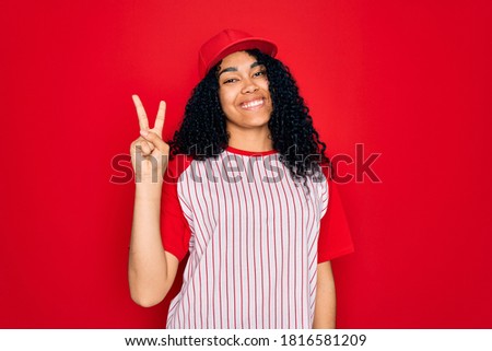 Young african american curly sportswoman wearing baseball cap and striped t-shirt showing and pointing up with fingers number two while smiling confident and happy.