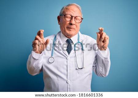Senior grey haired doctor man wearing stethoscope and medical coat over blue background gesturing finger crossed smiling with hope and eyes closed. Luck and superstitious concept.