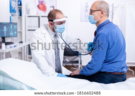 Doctor using stethoscope to listen senior man heart during examination in hospital room and wearing visor as safety precaution against coronavirus. Medical control for infections, disease. Royalty-Free Stock Photo #1816602488
