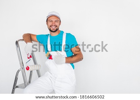 Smiling young male worker in uniform with roller and paintbrush showing thumb up gesture while standing on ladder against white wall during house renovation