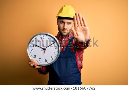 Young builder man wearing safety helmet holding big clock over yellow background with open hand doing stop sign with serious and confident expression, defense gesture