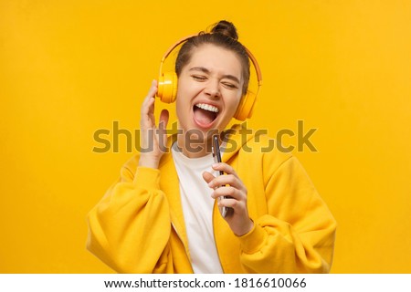Trendy young woman singing favorite song out loud in phone as mic, wearing wireless headphones, isolated on yellow background. Karaoke online app Royalty-Free Stock Photo #1816610066