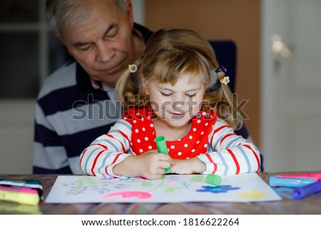 Cute little baby toddler girl and handsome senior grandfather painting with colorful felt pens and pencils at home. Grandchild and man having fun together, creative family.