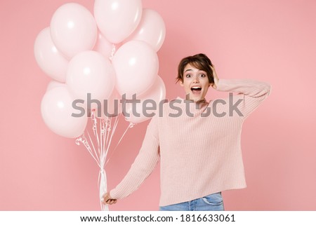 Surprised shocked happy young brunette woman in knitted casual sweater hold in hand air balloons put hand on head celebrating birthday holiday party isolated on pastel pink background studio portrait.